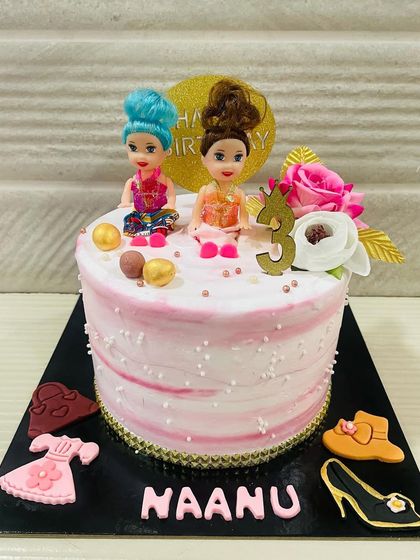A fun and fashionable Barbie-themed cake for a third birthday. This pink cake is decorated with two mini dolls and edible fondant accessories like dresses, hats, and shoes.