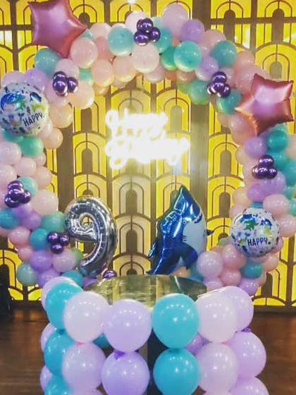 This balloon ring cake table is a unique and fun setup. The circular structure is decorated with pastel balloons and foil accents, with a 'Happy Birthday' neon sign glowing in the center.