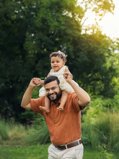 A father gives his daughter a shoulder ride during their outdoor family session. The lush green background and happy smiles make this a perfect portrait.