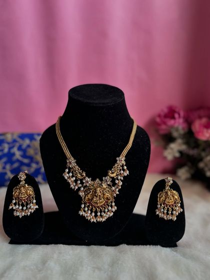 A simple and elegant temple jewellery necklace set. The centerpiece features a Goddess Lakshmi motif flanked by peacocks, with delicate pearl drops.