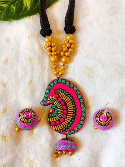A sleek necklace with a 'kalka' or paisley motif, here in a vibrant pink and green. The pendant has a small jhumka attached, and the set comes with matching earrings.