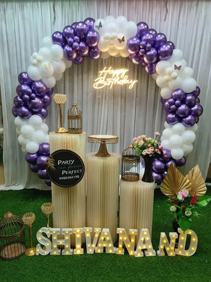 A lovely purple and white themed birthday decoration for Shivanand. The balloon ring is accented with butterfly details, and the setup includes a neon sign and the name in light-up letters on an artificial grass floor.
