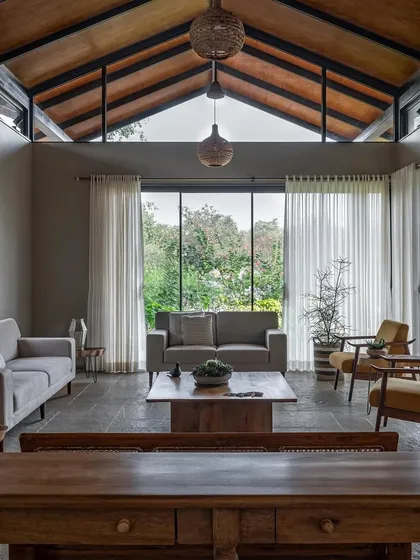 The living area of Grove House in Alibaug, designed as a gathering space for a veteran sailor and his family. The high, pitched roof and floor-to-ceiling glass walls dissolve the boundary between the interior and the surrounding coconut grove, creating a sanctuary filled with tropical light.