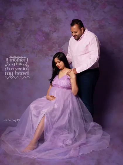 A gentle and loving portrait of a couple awaiting their baby. The father-to-be stands behind his partner, who is seated gracefully in a flowing purple gown, creating a protective and warm image.