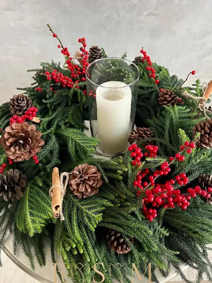 A closer look at this traditional Christmas wreath centerpiece. The pillar candle in the center casts a warm glow on the surrounding pine, berries, and cinnamon, creating a cozy holiday atmosphere.
