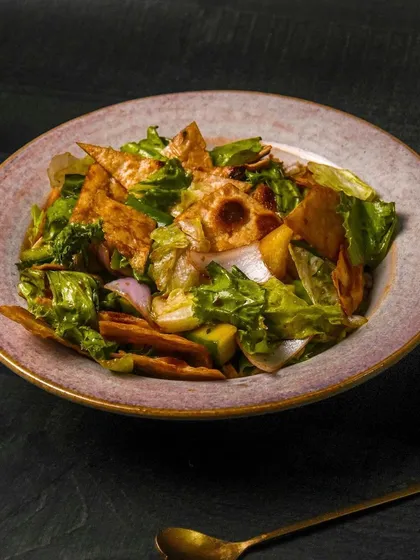 A fattoush salad with greens, vegetables, and crispy pita chips, tossed in a creamy dressing. The dark, rustic plate and background give this salad a gourmet look.