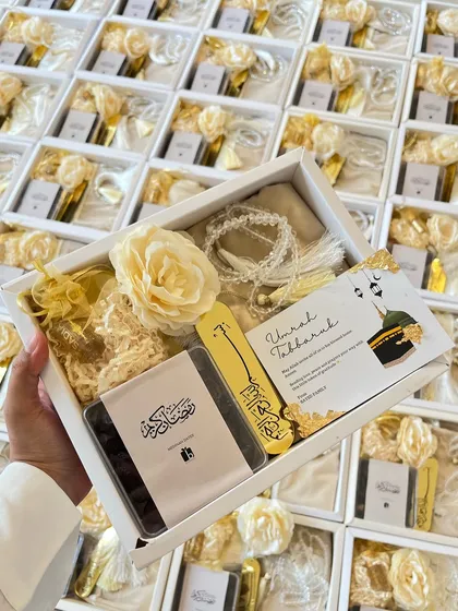 Holding up a completed Umrah return gift box, showing the beautiful arrangement of the prayer mat, Tasbeeh, bookmark, and other tokens of love.