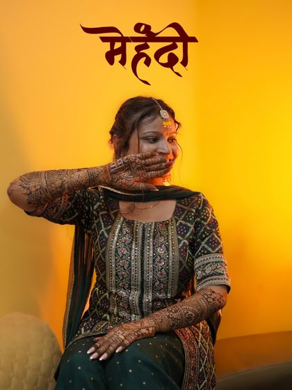 A playful and candid Mehendi portrait. The bride shyly covers her smile, drawing attention to the detailed henna art on her hands, set against a warm, glowing background.