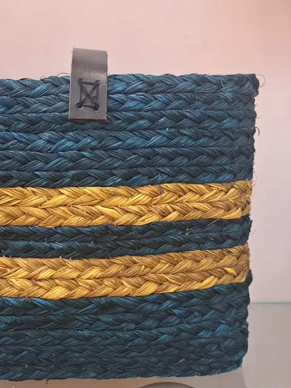A close-up of a rectangular woven basket in a deep teal color with two golden-yellow stripes. The leather tab detail adds a touch of sophistication, making it a chic storage option for any modern home.