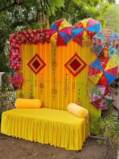 An outdoor Mehndi seating area with a backdrop of yellow drapes and colorful umbrellas. The traditional hangings and bright colors make it a fun and festive photo spot.