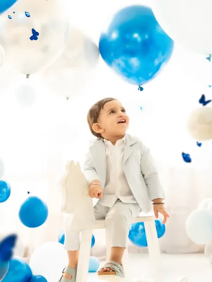 A baby boy on a rocking horse looks up in wonder at the surrounding balloons. This photo captures a moment of quiet curiosity and innocence during his first birthday celebration.