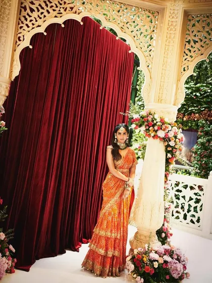 A vibrant and modern look for a wedding celebration. Here, Isha wears a stunning tiered orange saree by Arpita Mehta, bringing a bright pop of color to the festivities.