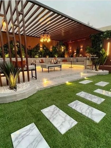A luxurious backyard designed for entertainment. This landscape features marble-style pavers set in artificial turf, a raised patio with a large seating area under a pergola, and dramatic uplighting on plants and structures.