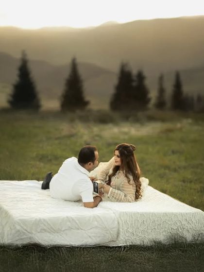 A surreal and romantic concept. The couple lies on a mattress in the middle of a vast, green meadow, with mountains in the background, creating a dreamy and unforgettable maternity portrait.