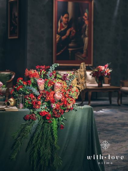 A grand floral installation that seems to flow from the table, set against the backdrop of a classic painting. The design is intentionally dramatic and abundant, echoing the opulence of the era.
