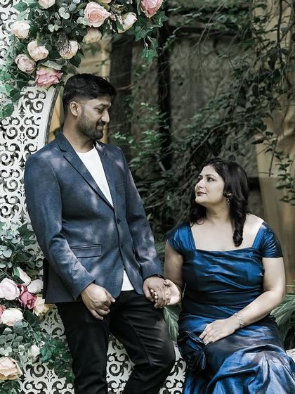 A couple holds hands while posing with a large, ornate crescent moon prop in a garden.