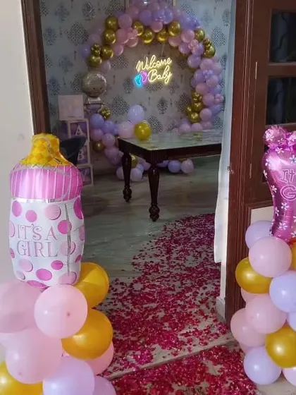 A sweet "Welcome Baby Girl" setup. The entrance features balloon pillars with "It's a Girl" foil balloons, leading to a room with a beautiful balloon arch and a "Welcome Baby" neon sign.