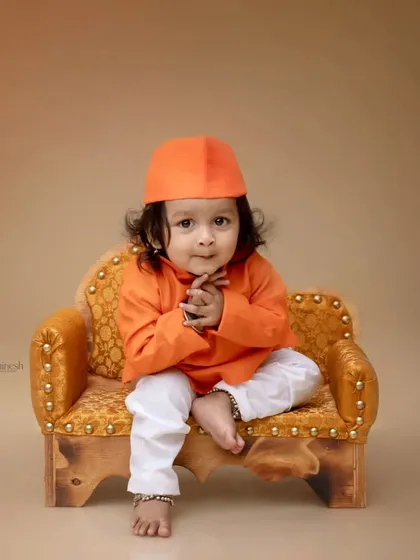 A charming portrait of a baby boy in a simple orange kurta, sitting on a miniature sofa. The neutral background ensures all attention is on his adorable pose.