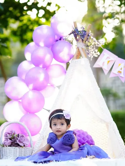An outdoor first birthday party setup with a teepee and purple balloons. The birthday girl looks adorable in her matching purple dress.