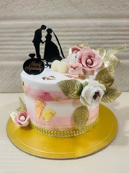 A romantic anniversary cake with a pink and white watercolor effect, adorned with golden leaves and a silhouette topper of a couple.