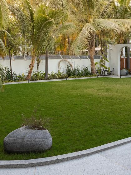 The expansive lawn of 'Tropical Manor', bordered by a clean granite edge and a natural stone planter. The design emphasizes open green space, framed by the arched wall and a grove of mature coconut palms.
