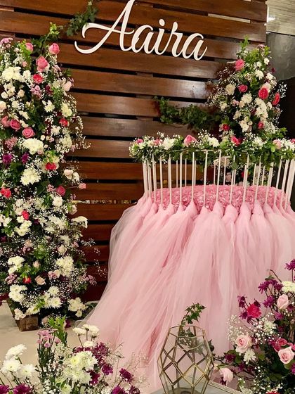 A beautiful shot of the cradle decor for baby Aaira's welcome party. The cradle is dressed in a pink tulle skirt and surrounded by towers of fresh flowers, with her name in a custom sign on the wooden backdrop.