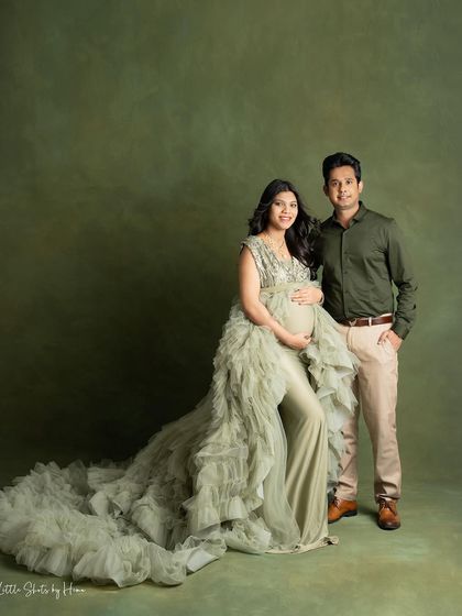 A stylish couple's portrait against an olive green backdrop. The mom-to-be wears a beautiful ruffled gown, standing with her partner for a classic and elegant photograph.