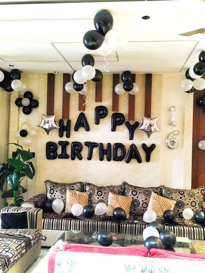 A simple yet elegant home decoration for a 16th birthday. The wall is decorated with "Happy Birthday" foil balloons in black, with clusters of black and white balloons floating on the ceiling.