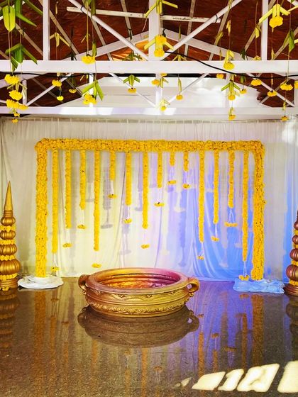 This indoor haldi setup features a backdrop of hanging marigold strings against a white curtain. The space is defined by traditional brass pots and a central urlu bowl for the ceremony, all under a ceiling of hanging flowers.
