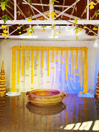 A complete setup for an outdoor Haldi ceremony. The backdrop features hanging marigold strings, and a traditional brass urli is placed in the center for the rituals, all framed by decorative golden pillars.