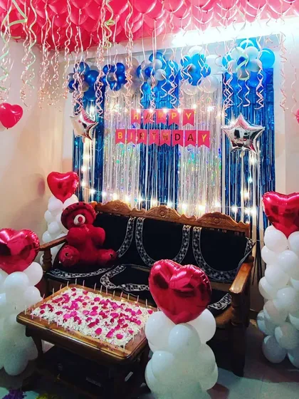 A colorful living room birthday setup using red, blue, and white balloons. The decor features a foil curtain backdrop, a teddy bear, and a table decorated with rose petals.
