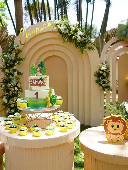 The beautiful cake and dessert station for Charvik's safari-themed first birthday. The setup includes a tiered cake, matching cupcakes, and is perfectly integrated with the modern, neutral-toned backdrop and animal props.
