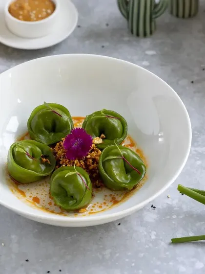 This is an example of modern, artistic food plating. The vibrant green of the spinach dumplings pops against the clean white bowl, while the careful placement of garnishes highlights the dish's elegance for a fine-dining menu.