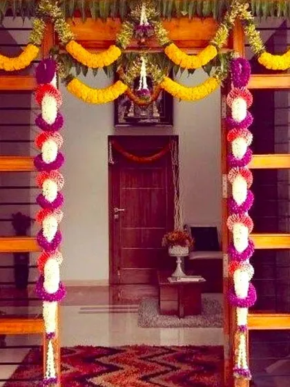 An elegant housewarming flower decoration for a main door. The wooden door is framed with colorful garlands of marigold, jasmine, and purple flowers.