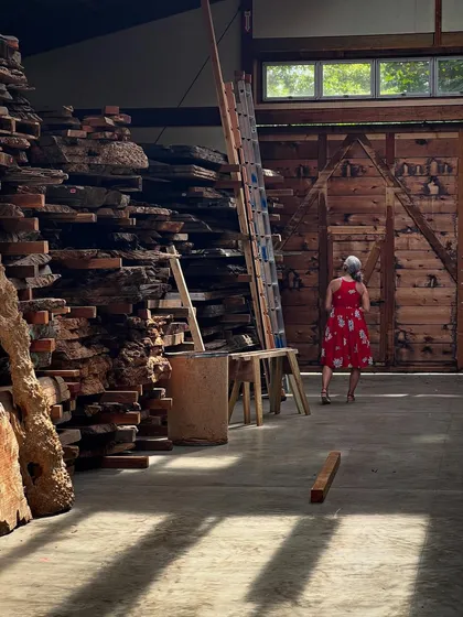 A woman in a red dress walks through the Nakashima wood storage building, surrounded by massive slabs of timber. The scale of the raw material is awe-inspiring, a reminder of the living trees from which it came.