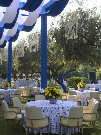 A Mediterranean-inspired haldi setup with blue and white drapes. The round tables with printed tablecloths and yellow floral centerpieces create a fresh and cheerful ambiance.