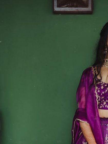 A bride in a vibrant purple lehenga, her smile lighting up the frame, stands next to a stone Buddha statue.
