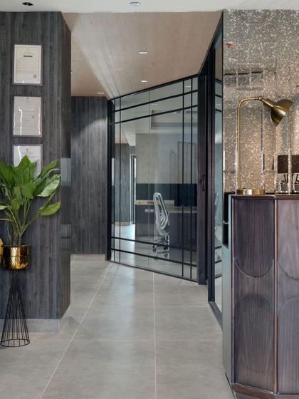 A view of the office corridor, showcasing the material palette of black ash wood, grey floor tiles, and terrazzo. The glass pivot door provides a glimpse into the workspace, maintaining a sense of transparency.