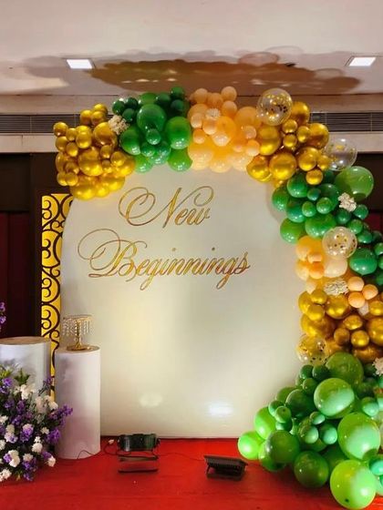 A sophisticated backdrop for an engagement party or roka ceremony. This setup features a 'New Beginnings' message on a white circular board, framed by an organic balloon arch in shades of green and gold.