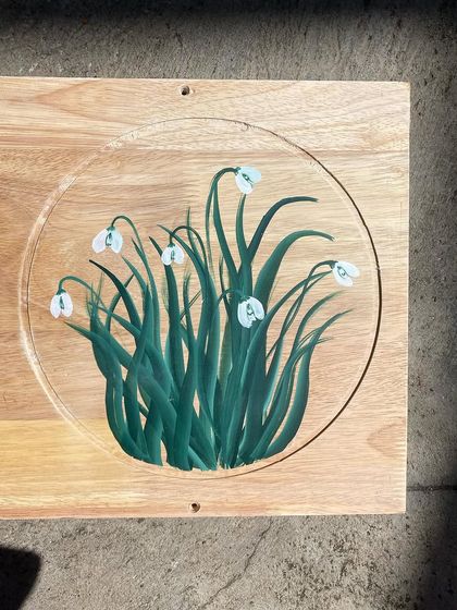 A cluster of snowdrop flowers, with their distinct bell shape, painted within the circular frame of the wooden panel.