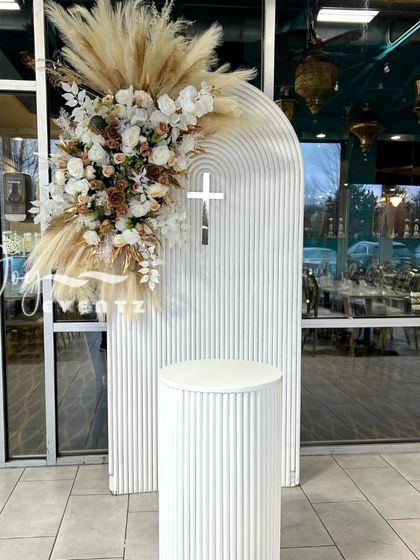 A simple and elegant christening decoration featuring a modern white arched panel with a cross. The arrangement is enhanced with a beautiful bouquet of pampas grass and white roses.