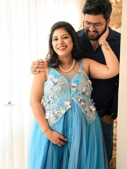 A bright, happy portrait of the couple by the window. The natural light illuminates their joyful expressions and the beautiful details of the blue maternity gown.