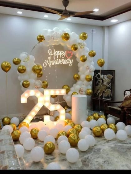 A beautiful 25th-anniversary setup with a white and gold balloon arch and a 'Happy Anniversary' neon sign.