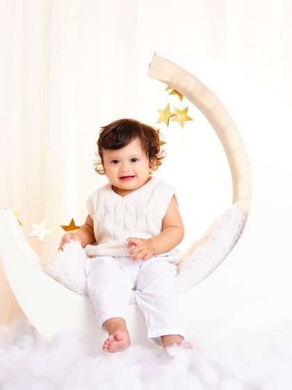 An adorable baby boy in a knitted vest, smiling for the camera while sitting on the crescent moon prop.