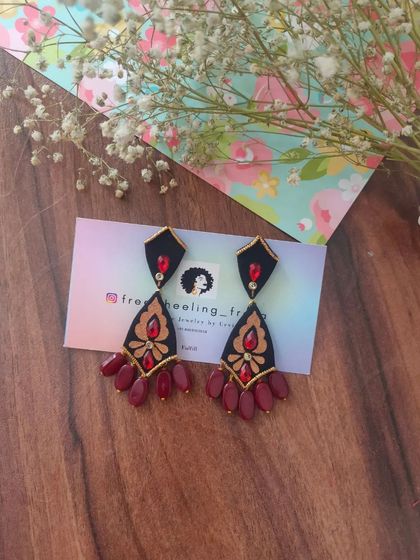A pair of elegant, hand-painted chandelier earrings. The black fabric base is adorned with golden and red painted details and finished with maroon bead drops.