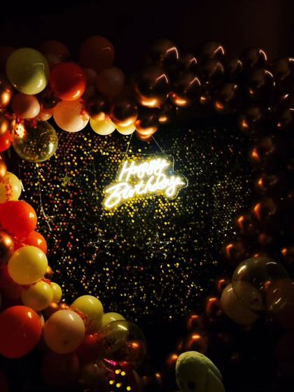 A classic birthday setup featuring a heart-shaped balloon arch and a 'Happy Birthday' neon sign against a sparkling sequin backdrop. The dark, moody lighting makes the colors and lights pop.