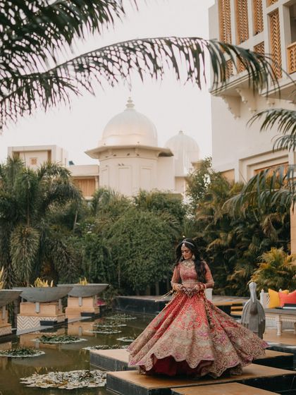 The bride stands gracefully by a lily pond at The Leela Palace Udaipur. The palace architecture and serene water provide a majestic backdrop for her magnificent lehenga.