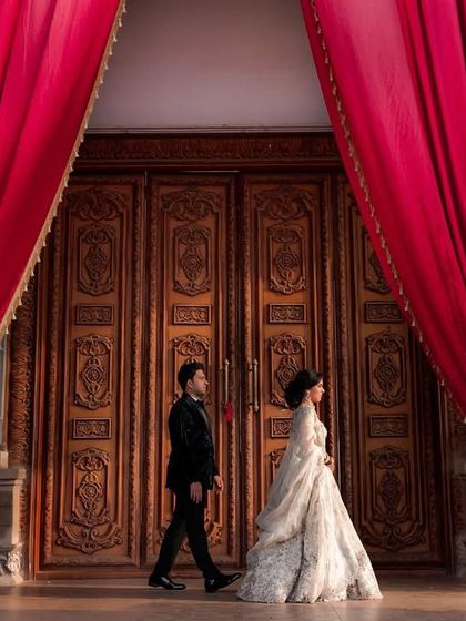 A grand, cinematic shot of the couple walking past ornate doors and dramatic red curtains. This image feels like a still from a classic movie, highlighting the elegance of their reception.