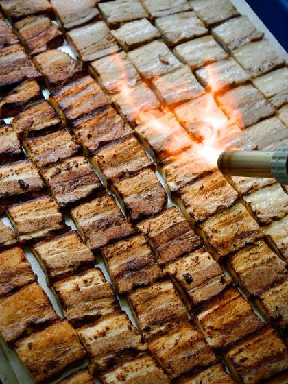 A chef uses a blowtorch to add the final char to rows of seasoned pork belly. This action shot captures the drama and precision of our live grilling stations, promising intense flavor.