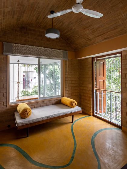 This serene seating area features exposed brick walls, a vaulted brick ceiling, and a vibrant yellow oxide floor with a flowing green inlay. The space opens to a small balcony, connecting the interior with the outdoors and ensuring cross-ventilation.